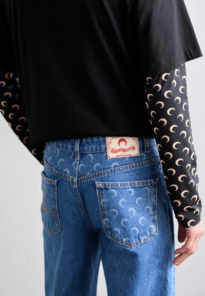 Svart långärmad skjorta med mönster av halvmånar, ihop med blå denimjeans med ett liknande subtilt tryck och en märkeslapp på midjebandet.