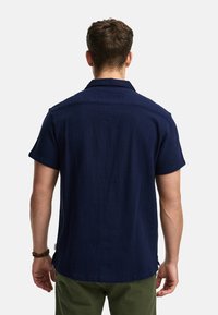 Camicia blu navy a maniche corte con colletto, tessuto strutturato e orlo curvo. Cuciture visibili e un'etichetta sul lato.