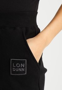 Black ribbed jogger pants feature a high waistband, side pockets, and a rectangular logo patch with "LON DUNN" in gray embroidery.