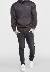 Sudadera negra con bolsillo frontal y logo, combinada con jeans oscuros y zapatillas blancas. Presenta un corte relajado y una textura suave.