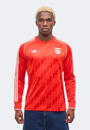 Young man with short curly blonde hair wearing a red long-sleeve sports jersey with white stripes and a team crest on the chest.