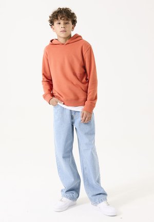 Hoodie orange avec une poche kangourou, associé à un jean baggy bleu clair et des baskets blanches, se tenant sur un fond neutre.