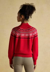Red knitted sweater with high ribbed collar, featuring white and black geometric patterns on the upper portion. Paired with houndstooth trousers.