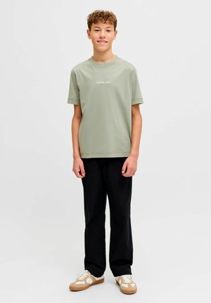 Teenager-Junge steht, trägt ein hellgrünes T-Shirt mit weißem Schriftzug, schwarze Hose und beige-weiße Turnschuhe, vor einem schlichten hellen Hintergrund.