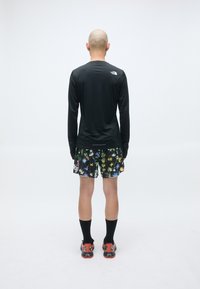 Schwarzes Langarmshirt, lebhaft gemusterte Shorts mit Blumenmuster, schwarze Socken und rote Sportschuhe; glatte Textur und enganliegendes Design.