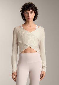 Világos bézs, bordázott, hosszú ujjú, V nyakú pakolós felső, amelyre egy halvány rózsaszín edző crop top és magas derekú leggings van rétegezve.