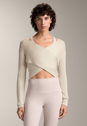 Hellbeige, geripptes Wickeloberteil mit langen Ärmeln und V-Ausschnitt, über einem blassrosa athletischen Crop-Top und hochtaillierten Leggings geschichtet.