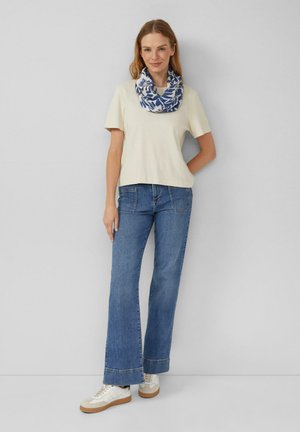 Frau trägt cremefarbenes Kurzarmoberteil, blauen Blumenschal, blaue Jeans und weiße Turnschuhe, steht vor einer schlichten hellgrauen Wand.