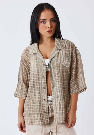 Vrouw met lang donker haar draagt een beige geruit overhemd over een witte sportbeha en lichtgekleurde broek, poseert tegen een witte achtergrond.