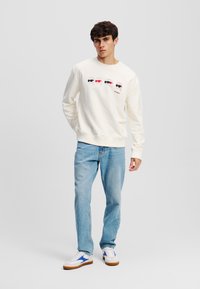 Crème sweatshirt met een grafisch ontwerp van vier gezichten die verschillende zonnebrillen dragen, gecombineerd met lichtblauwe jeans en witte sneakers met blauwe accenten.