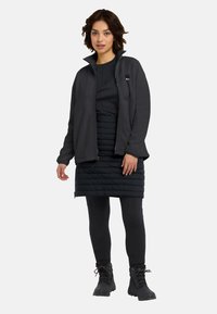 Giacca di pile nera con colletto alto, indossata sopra una gonna nera trapuntata. Il modello indossa anche leggings neri e scarponcini da trekking.