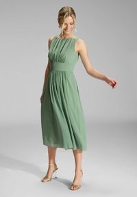 Robe midi sans manches verte avec une taille froncée et une jupe fluide. Le tissu a une texture légère ; portée avec des talons dorés à lanières.
