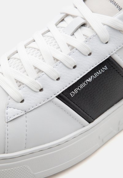 Baskets blanches avec lacets blancs, panneau noir affichant le logo Emporio Armani et semelle en caoutchouc texturée.