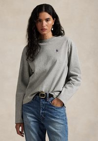 Kvinne med mørkt krøllete hår iført en grå langermet skjorte med logo og blå jeans, hånd i lommen, nøytral bakgrunn.