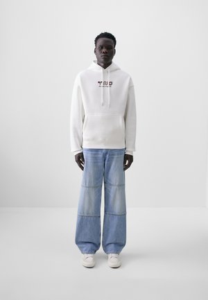 White hoodie with a front pocket, featuring colorful text. Light blue wide-leg jeans with panel stitching. White shoes. Minimal background.
