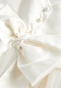 Gebroken wit satijnen stof, samengebracht in een grote strik met drie parelachtige knopen op een glad, vloeiend materiaal.