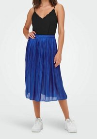 Sininen poimutettu midi-hame, jossa on tekstuuri, yhdistettynä mustaan topiin. Valkoiset lenkkarit viimeistelevät asun. Sujuva vyötäröyksityiskohta.