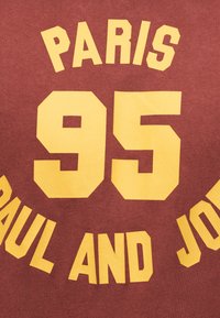 Μαρόν ύφασμα με μεγάλα κίτρινα γράμματα «PARIS 95 PAUL AND JOE» σε έντονους κεφαλαίους χαρακτήρες.