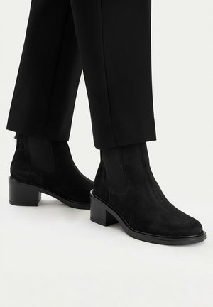Černé kotníkové boty z broušené kůže s blokovým podpatkem nošené s černými kalhotami s rovnými nohavicemi na neutrálním pozadí.