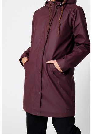 Personne portant un long imperméable à capuche violet foncé, les mains dans les poches avant, debout devant un fond clair uni.