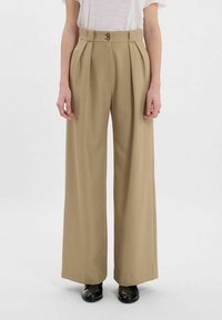 Pantalons larges beige avec des plis et une taille haute, dotés de deux boutons à l'avant, fabriqués en un tissu lisse. Associés à des chaussures noires.