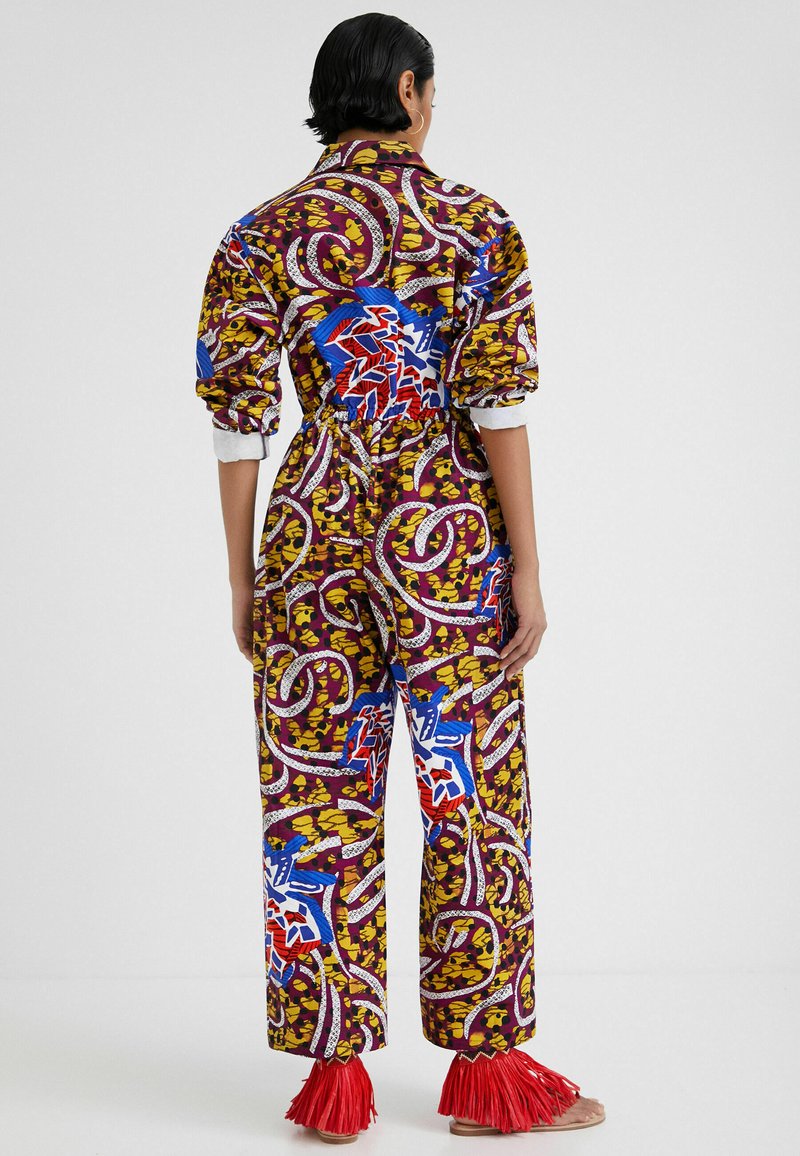 Mono multicolor con patrones abstractos llamativos en púrpura, azul y amarillo. Presenta una cintura elástica y acentos de flecos rojos en los tobillos.