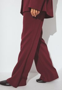 Burgundijas platās bikses, kas izgatavotas no mīkstas, teksturētas auduma ar skaidrām līnijām; pieskaņotas spīdīgām melnām kurpēm ar norādītu purngalu.