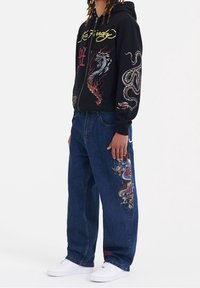 Svart huvtröja med dragkedja och livfullt broderi av drake och orm. Blå lösa jeans har en drake-design på benet och vita sneakers.