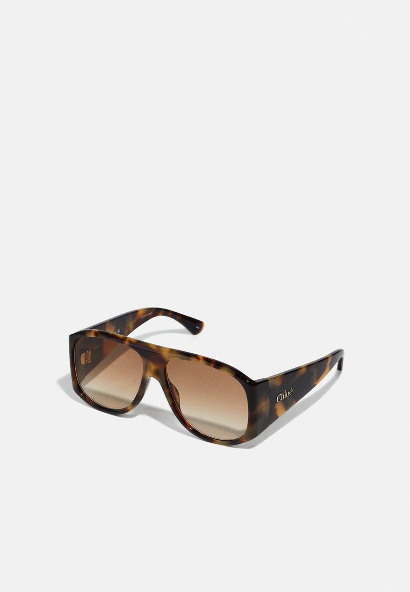 Masivní sluneční brýle ve tvaru želvoviny s hnědými gradientovými skly; obdélníkový tvar a ploché strany. Logo vyryté na nožce.