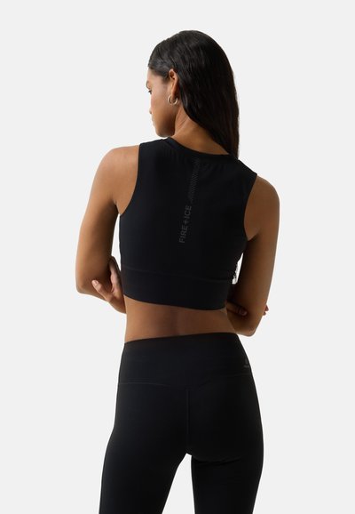 Frau mit langen dunklen Haaren, die ein schwarzes, ärmelloses Crop-Top und hoch taillierte Leggings trägt, steht mit verschränkten Armen und dem Rücken zur Kamera.