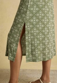 Falda verde con estampado y una abertura lateral que muestra las piernas de la modelo, combinada con sandalias planas blancas de tiras sobre un suelo de piedra.