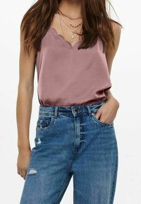 En kvinna modellerar en ärmlös topp med scallop-halsringning i mauve och högmidjade, slitna blå jeans, stående mot en neutral bakgrund.