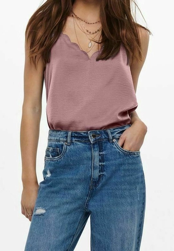 En kvinna modellerar en ärmlös topp med scallop-halsringning i mauve och högmidjade, slitna blå jeans, stående mot en neutral bakgrund.