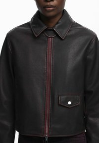 Giubbotto di pelle nera con colletto strutturato, zip frontale e taschino sul petto. Presenta dettagli di cuciture rosse e una texture liscia.