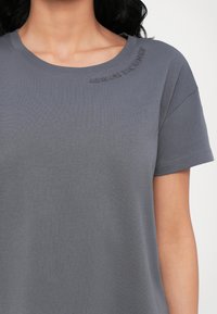 Grijze T-shirt met korte mouwen en een ronde halslijn. Kenmerkt zich door de verhoogde tekst "ARMANI EXCHANGE" nabij de kraag. Vloeibare, lichte katoenen stof.