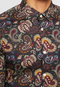 Nappikauluksinen paita, jossa on tumma tausta ja monimutkaiset paisley-kuviot punaisena, vihreänä ja keltaisena, valmistettu sileästä kankaasta.