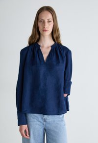 Blusa in lino blu navy con scollatura a V, dettagli di raccolto sul collo e maniche lunghe. Abbinata a jeans in denim azzurro.