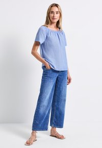 Top blu chiaro a maniche corte con scollo arricciato, abbinato a jeans in denim blu a gamba larga e sandali beige con fasce. Look semplice e casual.