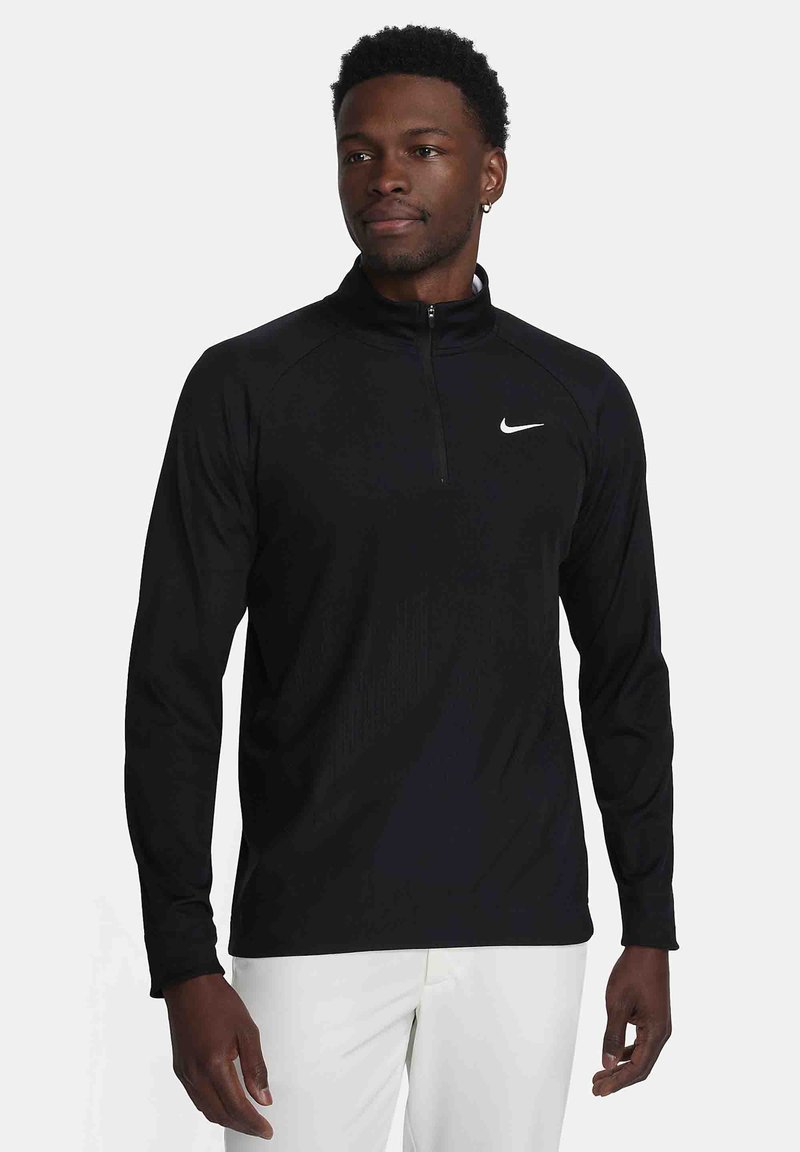 Moški oblečen v črno dolgosleeve pulover Nike z kratkim zadrgo in bele hlače, stoji pred enostavno svetlo ozadje.