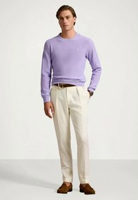 Ανοιχτό μωβ πουλόβερ με υφή πλεκτού, συνδυασμένο με κρέμα παντελόνι. Καφέ ζώνη και λουστρινό παπούτσι συμπληρώνουν το ντύσιμο. Ουδέτερο φόντο.