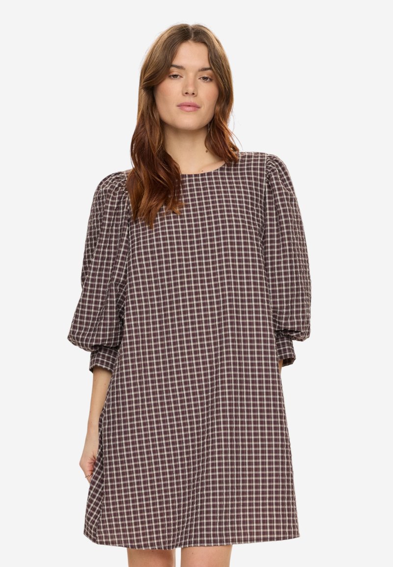 Femme aux cheveux bruns portant une robe ample mi-longue à carreaux violets et blancs avec des manches trois-quarts bouffantes.