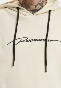 Felpa in cotone color crema con stringhe nere, con testo corsivo nero "Rocawear" sulla parte anteriore. Texture liscia, design casual.