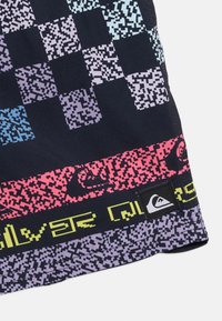 Κοντινή λήψη υφάσματος με πολύχρωμα, ψηφιακά σχεδιασμένα μοτίβα σε σκακιέρα και ρίγες, με ένα μικρό λογότυπο βουνού σε ασπρόμαυρο.