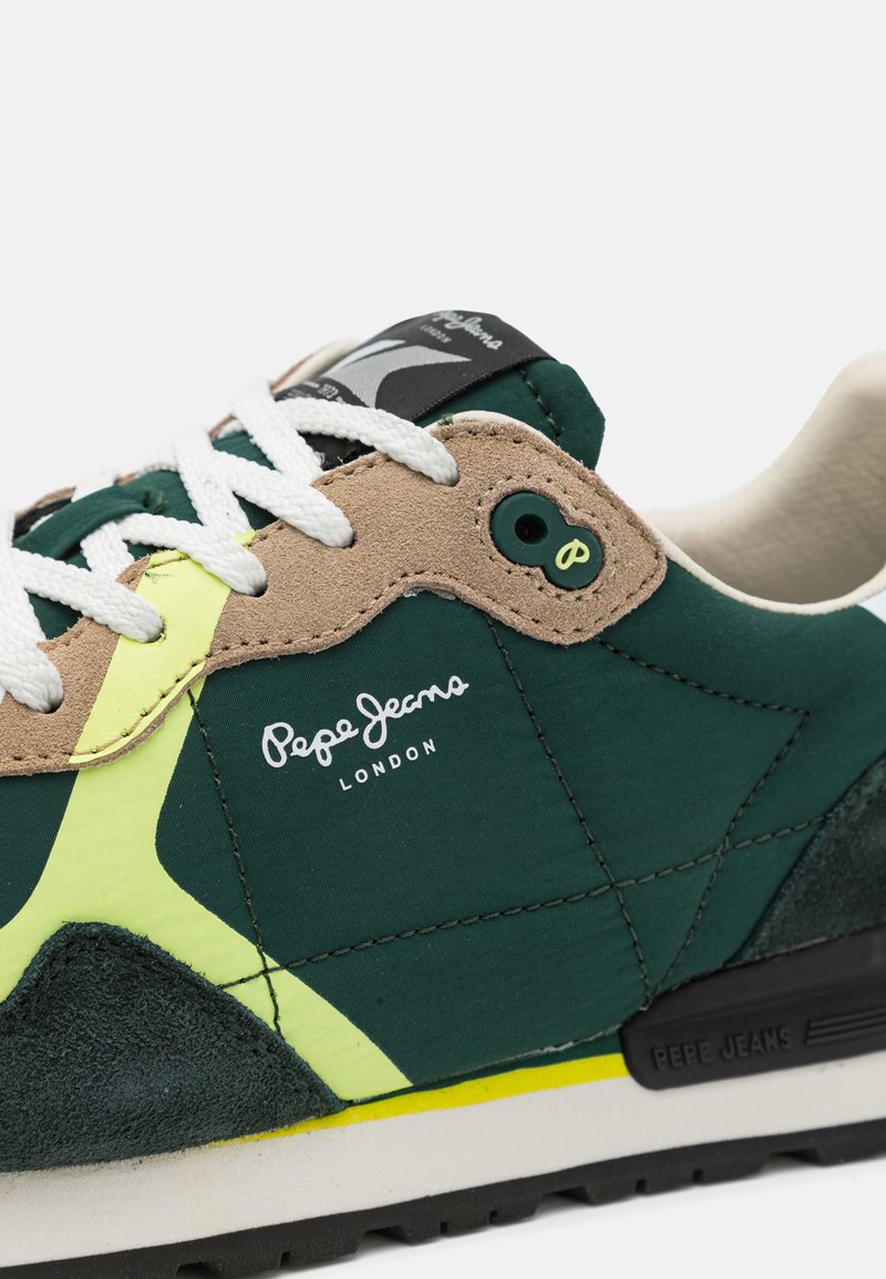 Zapatilla verde y beige con una parte superior texturizada, cordones blancos, acentos amarillos y una etiqueta negra con el logo "Pepe Jeans".