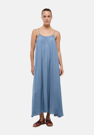 Femme debout portant une robe maxi bleue ample sans manches avec des bretelles fines et des sandales marron à brides sur un fond clair uni.