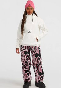 Woman with long braids wearing a pink beanie, white hooded sweatshirt, black patterned pants, and black boots standing against a plain background.