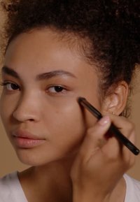 Modèle appliquant du maquillage avec un pinceau, mettant en valeur un teint naturel, une peau légèrement texturée et une forme de sourcils définie sur un fond neutre.