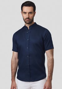 Niebieska koszula z krótkim rękawem i kołnierzykiem mandaryńskim, gładka w dotyku, z zapięciem na guziki z przodu, noszona z jasnymi spodniami.