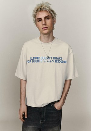 Giovane con capelli biondi spettinati che indossa una maglietta beige con il testo blu "LA VITA NON SI FERMA PER I DUBBI 2026" e jeans scuri.