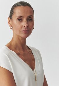 V-hihäntävalkoinen toppi lyhyillä hihoilla. Pitkä kultaketju riipuksella ja kultaiset renkaakorvaket. Sileä pinta, neutraali tausta.